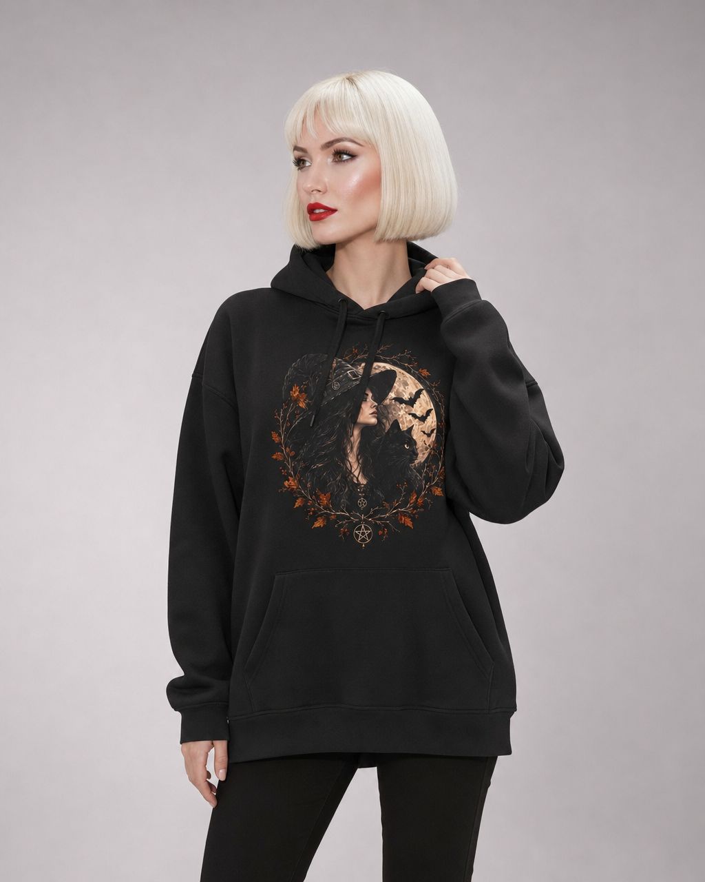Model with short platinum-blonde hair wearing a black hoodie with drawstring hood and kangaroo pocket. The chest features a gothic autumnal print: a long-haired woman with a black cat under a full moon, framed by fall leaves, branches, and bat silhouettes, with a pentagram at the bottom. 