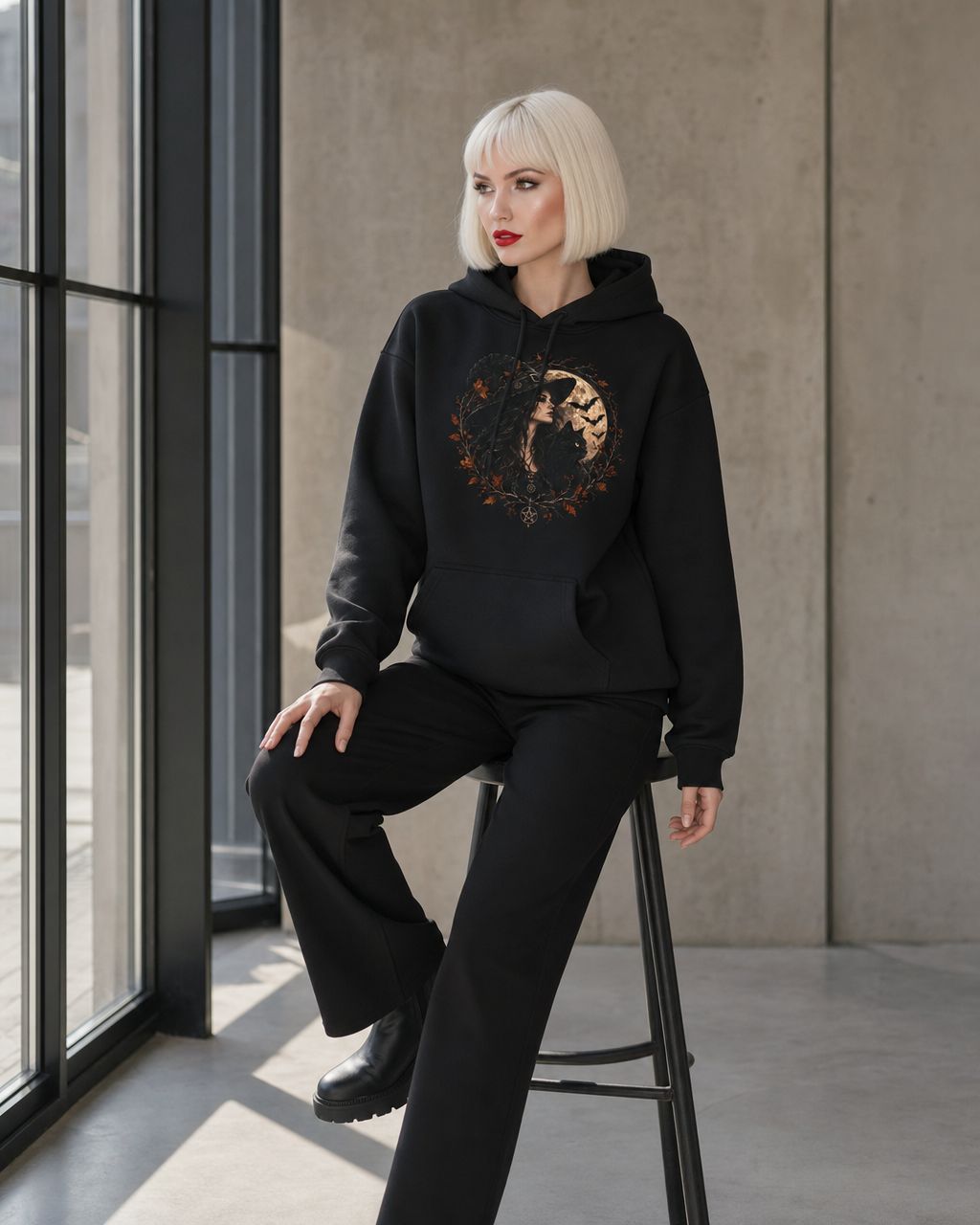 Model with short platinum-blonde hair wearing a black hoodie with drawstring hood and kangaroo pocket. The chest features a gothic autumnal print: a long-haired woman with a black cat under a full moon, framed by fall leaves, branches, and bat silhouettes, with a pentagram at the bottom. 